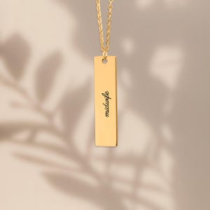 May include: A gold-toned vertical bar pendant necklace featuring a delicate chain and a polished rectangular metal tag. The word midwife is engraved in a black cursive script font centered on the front surface of the gold bar pendant.