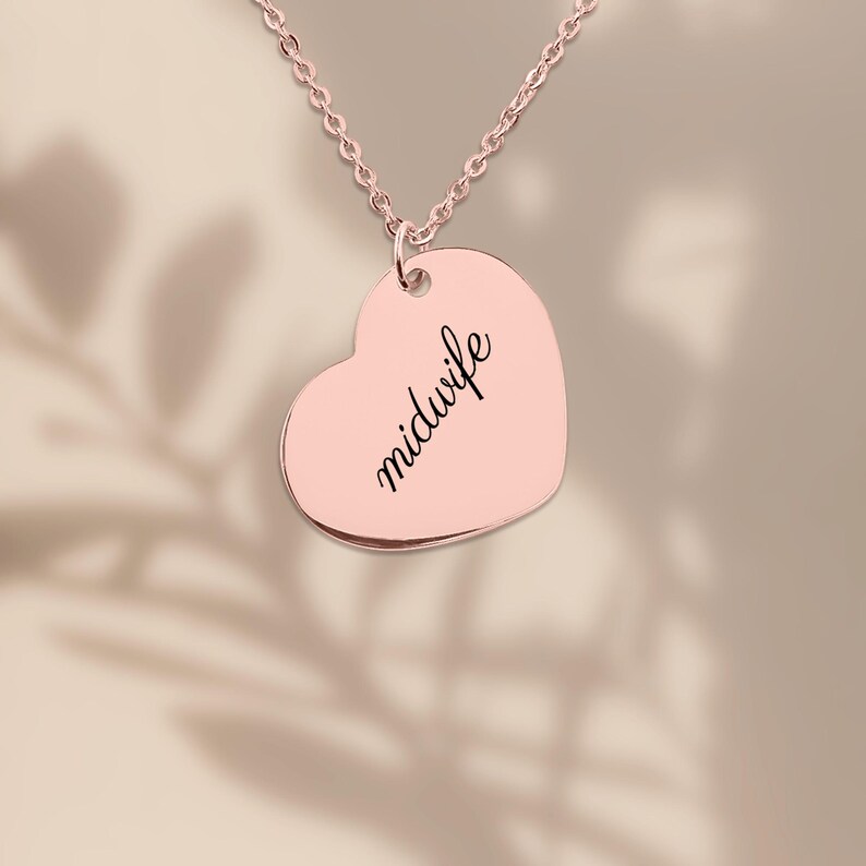 May include: A rose gold heart-shaped pendant necklace featuring the word midwife engraved in a black cursive script. The pendant hangs from a delicate matching rose gold chain, creating a simple and polished piece of jewelry.