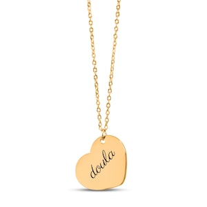 Puede incluir: Un collar de tono dorado con una cadena fina y un colgante en forma de coraz&oacute;n. El colgante tiene grabado el texto "doula" en una fuente cursiva de color negro. La pieza cuelga verticalmente, mostrando la superficie lisa y pulida del coraz&oacute;n.