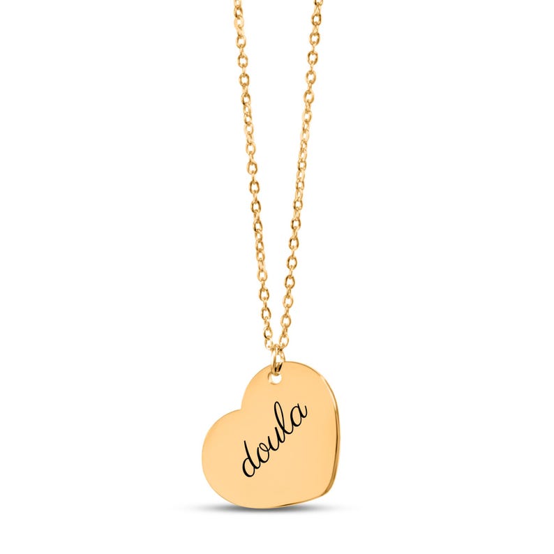 Puede incluir: Un collar de tono dorado con una cadena fina y un colgante en forma de coraz&oacute;n. El colgante tiene grabado el texto "doula" en una fuente cursiva de color negro. La pieza cuelga verticalmente, mostrando la superficie lisa y pulida del coraz&oacute;n.