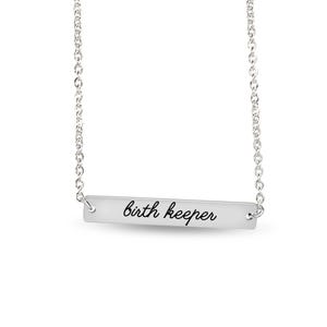 Puede incluir: Un collar de barra horizontal plateada con una cadena fina y un colgante rectangular. El colgante tiene grabado el texto "birth keeper" en una fuente cursiva negra, centrado sobre la superficie met&aacute;lica lisa del accesorio.
