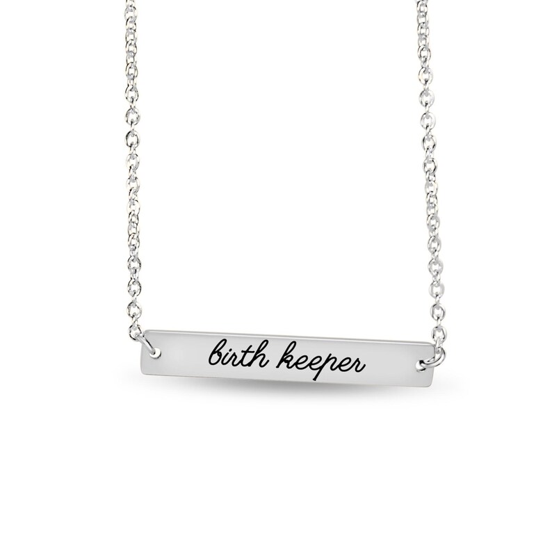 Puede incluir: Un collar de barra horizontal plateada con una cadena fina y un colgante rectangular. El colgante tiene grabado el texto "birth keeper" en una fuente cursiva negra, centrado sobre la superficie met&aacute;lica lisa del accesorio.