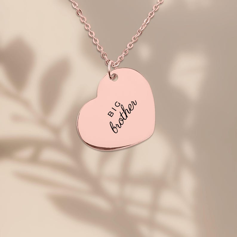 Op de afbeelding: Een hartvormige hanger aan een ketting in ros&eacute;goud met de gegraveerde tekst "BIG brother" in een combinatie van een zwart schreefloos lettertype en een scriptlettertype. De hanger hangt aan een bijpassende fijne ketting.