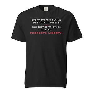 Puede incluir: Una camiseta negra de manga corta con texto en blanco que dice: EVERY SYSTEM CLAIMS TO PROTECT SAFETY. THE TEST IS WHETHER IT ALSO PROTECTS LIBERTY. La palabra LIBERTY está impresa en rojo, con una línea horizontal roja encima.