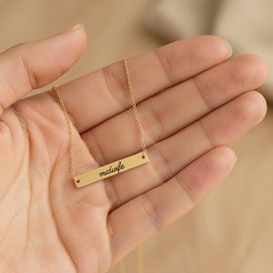 May include: A gold-toned bar necklace featuring a delicate chain and a horizontal rectangular pendant. The pendant is engraved with the word midwife in a black cursive script font, centered on the smooth, polished metal surface.