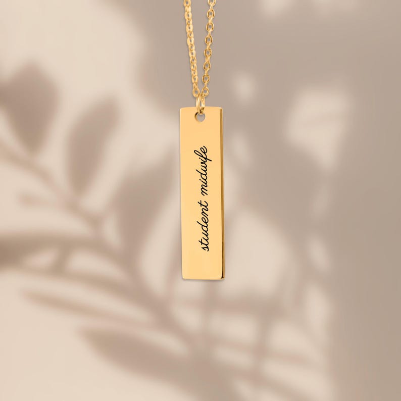 Puede incluir: Un collar con colgante de barra vertical en tono dorado con una cadena delicada y un dije rectangular. El dije est&aacute; grabado con el texto negro student midwife en una fuente cursiva, centrado verticalmente en la superficie met&aacute;lica lisa.
