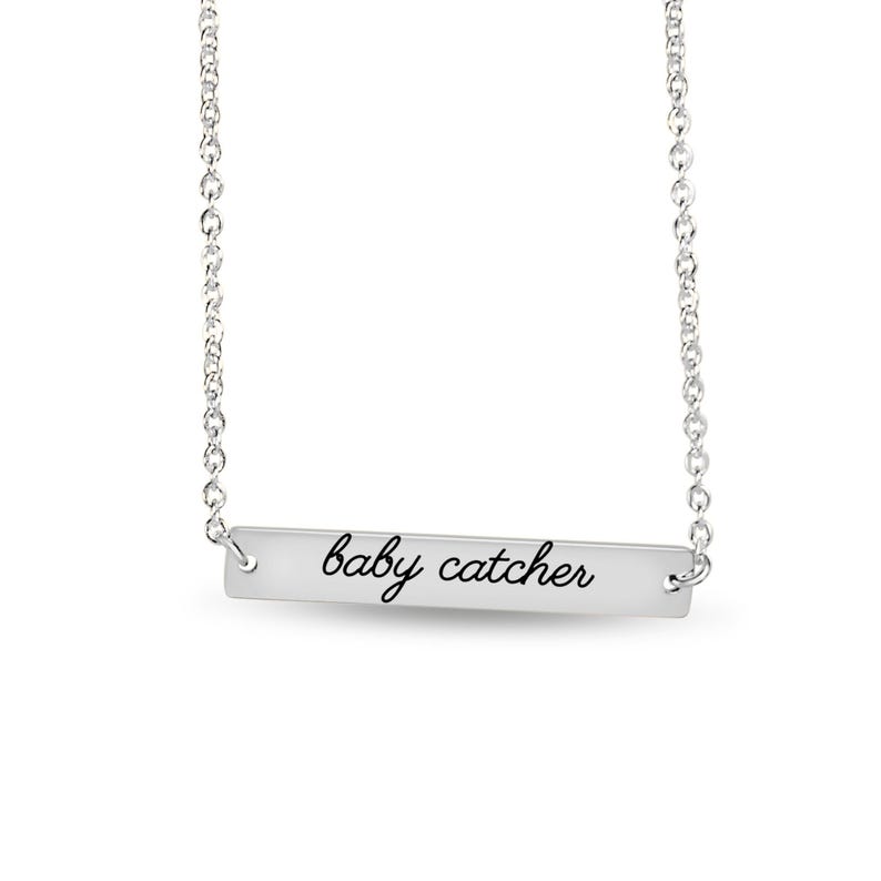 May include: A silver-toned horizontal bar necklace featuring a delicate chain and a rectangular metal pendant. The pendant is engraved with the text "baby catcher" in a black, cursive script font, centered on the polished metallic surface.
