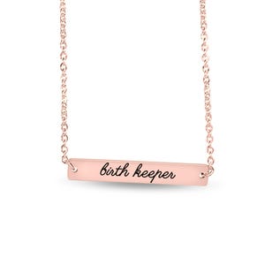 Puede incluir: Un collar de barra en tono oro rosa con una cadena fina y un colgante rectangular horizontal. El colgante tiene grabado en negro y en cursiva el texto birth keeper, centrado sobre la superficie met&aacute;lica lisa y pulida.