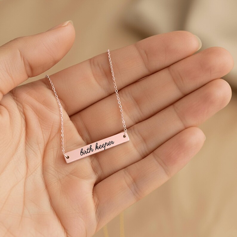 Puede incluir: Un collar de barra en tono oro rosa con una cadena delicada y un colgante rectangular horizontal. El colgante est&aacute; grabado con el texto "birth keeper" en una fuente cursiva negra, centrado en la superficie met&aacute;lica.