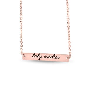 May include: A rose gold bar necklace featuring a delicate chain and a horizontal rectangular pendant. The pendant is engraved with the text "baby catcher" in a black cursive script font, centered on the polished metal surface.
