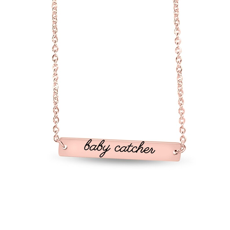 May include: A rose gold bar necklace featuring a delicate chain and a horizontal rectangular pendant. The pendant is engraved with the text "baby catcher" in a black cursive script font, centered on the polished metal surface.