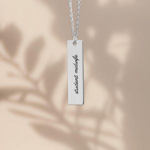 Puede incluir: Un collar con colgante de barra vertical plateado que presenta una cadena delicada y una placa rectangular de metal pulido. El colgante est&aacute; grabado con el texto cursivo negro student midwife, posicionado verticalmente a lo largo del centro.