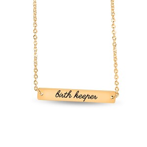 Puede incluir: Un collar de barra de metal dorado con una cadena fina y un colgante rectangular horizontal. El colgante tiene grabado el texto "birth keeper" en cursiva negra centrado sobre su superficie lisa y pulida.