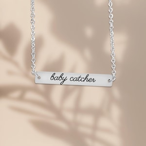 May include: A silver-toned horizontal bar necklace featuring a delicate chain and a rectangular metal pendant. The pendant is engraved with the text baby catcher in a black cursive script font, centered on the smooth metallic surface.