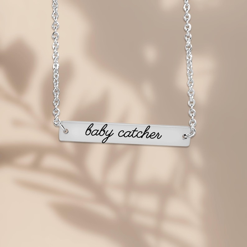 May include: A silver-toned horizontal bar necklace featuring a delicate chain and a rectangular metal pendant. The pendant is engraved with the text baby catcher in a black cursive script font, centered on the smooth metallic surface.