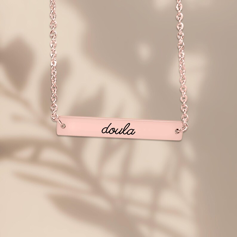 Pode incluir: Um colar de barra em ouro rosa com uma corrente delicada e um pingente retangular horizontal. O centro da barra est&aacute; gravado com a palavra doula em uma fonte cursiva preta, suspenso por dois pequenos an&eacute;is de jun&ccedil;&atilde;o circulares.