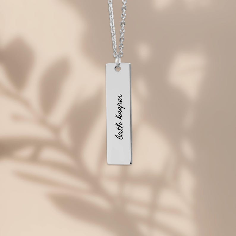 Op de afbeelding: Een zilverkleurige ketting met een verticale staafhanger, bestaande uit een fijne ketting en een gepolijst rechthoekig metalen plaatje. De hanger is gegraveerd met de zwarte tekst Birth keeper, verticaal in het midden van de staaf.