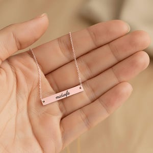 May include: A rose gold-toned bar necklace featuring a delicate chain and a horizontal rectangular pendant. The pendant is engraved with the word midwife in a black cursive script font, centered on the smooth, polished metal surface.