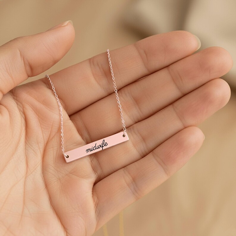 May include: A rose gold-toned bar necklace featuring a delicate chain and a horizontal rectangular pendant. The pendant is engraved with the word midwife in a black cursive script font, centered on the smooth, polished metal surface.