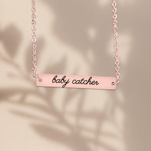 May include: A rose gold bar necklace featuring a delicate chain and a horizontal rectangular pendant. The pendant is engraved with the text baby catcher in a black cursive script font, centered on the polished rose gold surface.