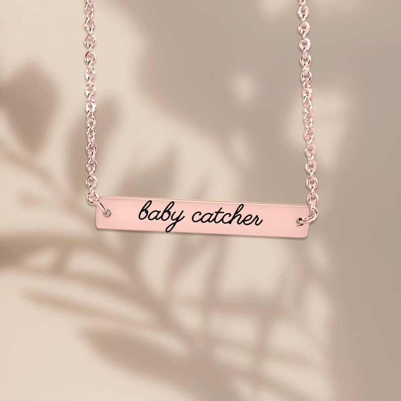May include: A rose gold bar necklace featuring a delicate chain and a horizontal rectangular pendant. The pendant is engraved with the text baby catcher in a black cursive script font, centered on the polished rose gold surface.