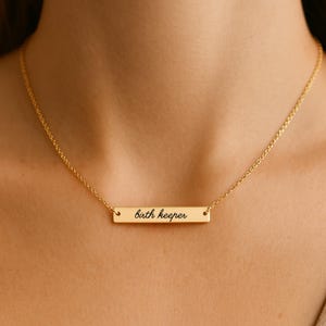 Puede incluir: Un collar de tono dorado con un colgante de barra rectangular horizontal. La barra est&aacute; grabada con el texto "birth keeper" en una fuente cursiva negra. El colgante cuelga de una delicada cadena de tono dorado alrededor del cuello.