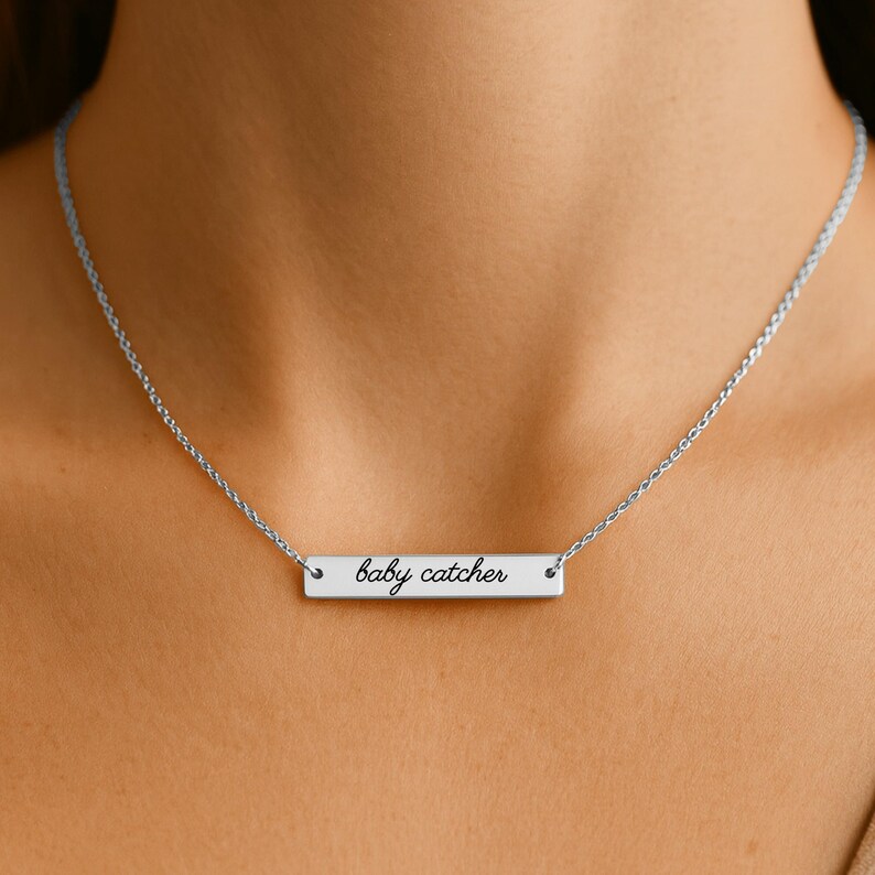May include: A silver-toned horizontal bar necklace featuring the text "baby catcher" engraved in a black cursive script. The pendant is suspended from a delicate, thin silver-toned chain that rests around the neck, showcasing a polished metallic finish.