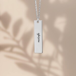May include: A silver-toned vertical bar pendant necklace featuring the word midwife engraved in a black script font. The pendant hangs from a matching silver-toned chain, creating a clean and simple piece of jewelry with a polished metallic finish.