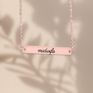 May include: A rose gold-toned bar necklace featuring a delicate chain and a horizontal rectangular pendant. The pendant is engraved with the word midwife in a black cursive script font, centered on the smooth metallic surface.