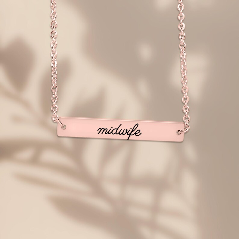 May include: A rose gold-toned bar necklace featuring a delicate chain and a horizontal rectangular pendant. The pendant is engraved with the word midwife in a black cursive script font, centered on the smooth metallic surface.