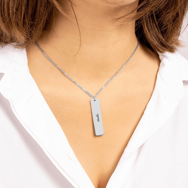 May include: A silver-toned vertical bar pendant necklace featuring a delicate chain and a rectangular metal tag engraved with the word doula in a cursive script font, suspended from a simple link chain that rests against the collarbone.