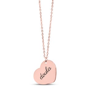 Puede incluir: Un collar con colgante en forma de coraz&oacute;n de color oro rosa que presenta la palabra doula grabada en una tipograf&iacute;a cursiva negra. El colgante cuelga de una delicada cadena a juego de color oro rosa con peque&ntilde;os eslabones ovalados, creando una joya sencilla.