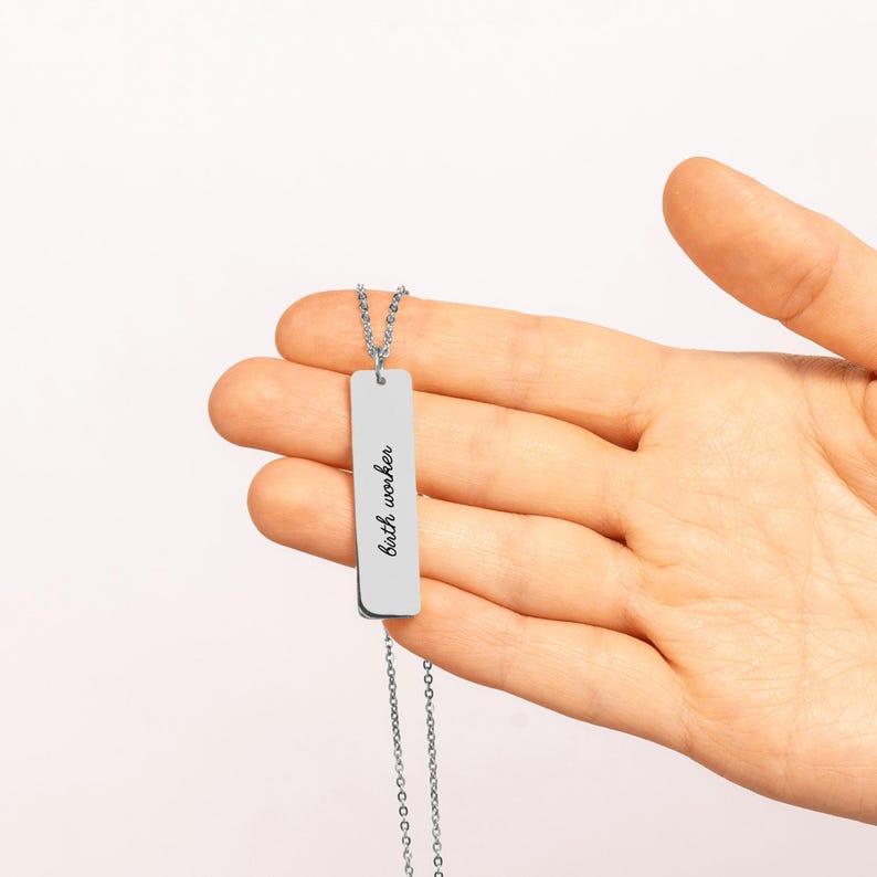 May include: A silver-toned vertical bar pendant necklace featuring the engraved text "birth worker" in a cursive script font. The rectangular metal charm hangs from a delicate matching silver-toned chain, displaying a clean and polished finish.