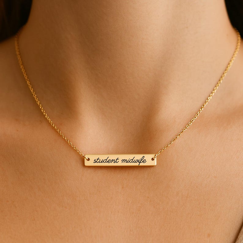 May include: A gold-toned bar necklace featuring a delicate chain and a horizontal rectangular pendant. The pendant is engraved with the text student midwife in a black cursive script font, centered on the polished metal surface.