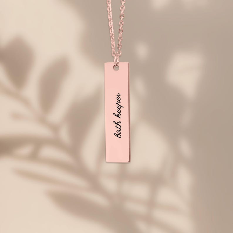 Op de afbeelding: Een ros&eacute;goudkleurige verticale staafhanger ketting met een fijne ketting en een rechthoekig metalen plaatje gegraveerd met de zwarte scripttekst "birth keeper" gecentreerd op de voorzijde van de gladde, gepolijste metalen staaf.