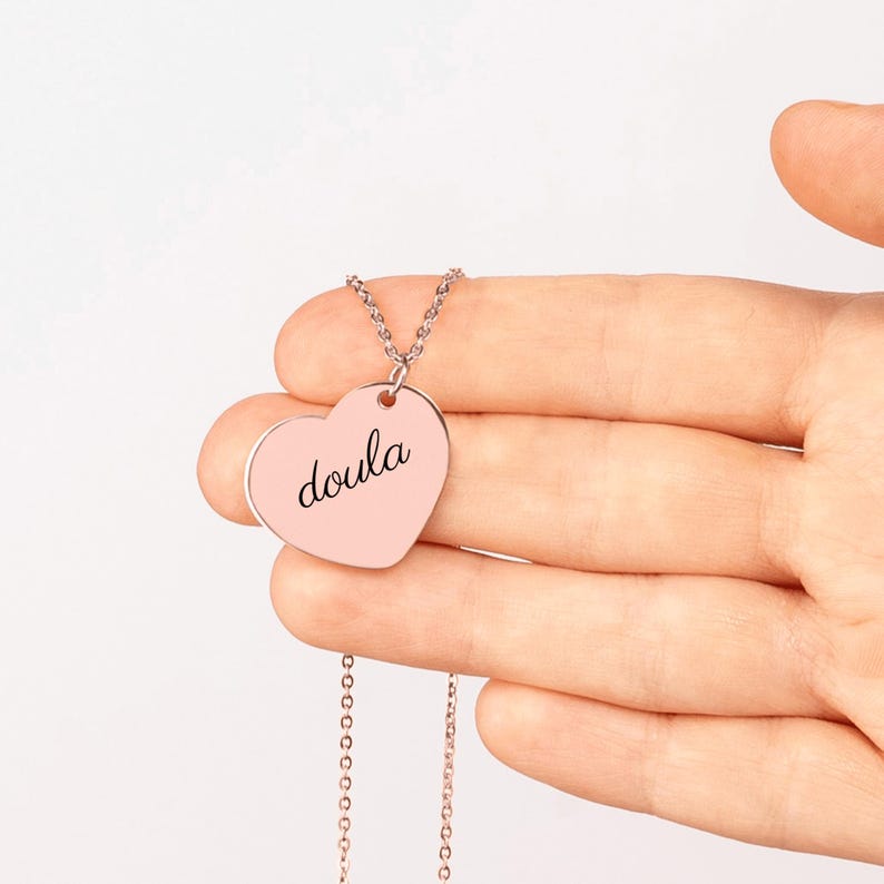 Puede incluir: Un collar con colgante en forma de coraz&oacute;n en oro rosa que presenta la palabra doula grabada en una tipograf&iacute;a cursiva negra. El colgante est&aacute; unido a una delicada cadena a juego en oro rosa, creando una pieza de joyer&iacute;a sencilla.