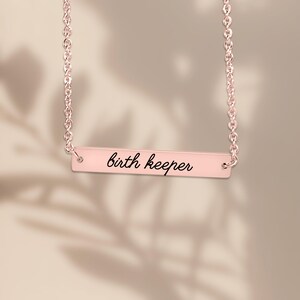 Puede incluir: Un collar de barra en oro rosa con una cadena delicada y un colgante rectangular horizontal. El colgante est&aacute; grabado con el texto en cursiva negra birth keeper, centrado en la superficie met&aacute;lica pulida de la barra.