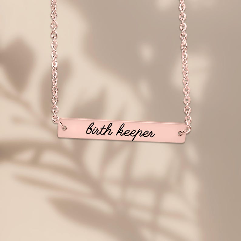 Puede incluir: Un collar de barra en oro rosa con una cadena delicada y un colgante rectangular horizontal. El colgante est&aacute; grabado con el texto en cursiva negra birth keeper, centrado en la superficie met&aacute;lica pulida de la barra.
