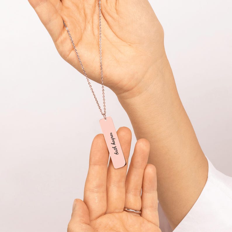 Op de afbeelding: Een zilverkleurige ketting met een verticale, rechthoekige lichtroze hanger. De hanger is gegraveerd met de tekst "birth keeper" in een zwart cursief lettertype. De ketting is bevestigd aan een dunne, verfijnde metalen schakelketting.