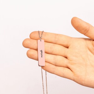 May include: A rose gold-toned vertical bar pendant necklace featuring the word midwife engraved in a black cursive script. The rectangular metal bar hangs from a delicate matching chain, creating a simple and elegant piece of jewelry.