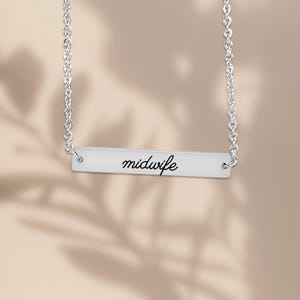 May include: A silver-toned horizontal bar necklace featuring the word midwife engraved in a black cursive script. The pendant is suspended from a delicate silver-toned chain with circular links, creating a clean and simple jewelry piece.