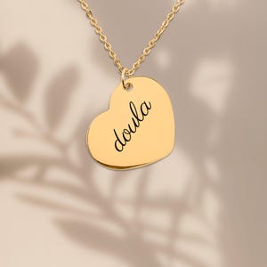 Puede incluir: Un collar con colgante en forma de coraz&oacute;n de tono dorado que presenta la palabra doula grabada en una tipograf&iacute;a cursiva negra. El colgante cuelga de una delicada cadena dorada a juego, creando una pieza de joyer&iacute;a sencilla.