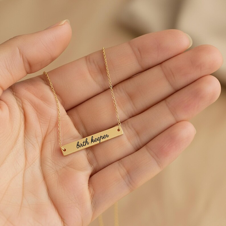 Puede incluir: Un collar de barra en tono dorado con una cadena fina y un colgante rectangular horizontal. El colgante tiene grabado el texto "birth keeper" en una fuente cursiva negra, centrado sobre la superficie de metal pulido.