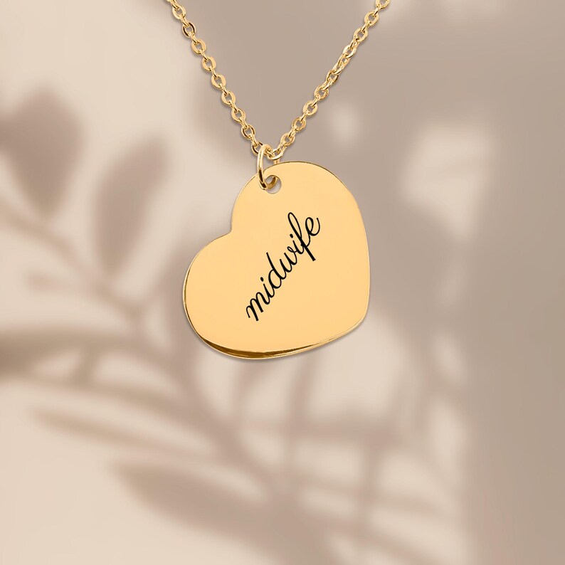 May include: A gold-toned heart-shaped pendant necklace featuring the word midwife engraved in a black cursive script. The pendant hangs from a delicate gold-toned chain, showcasing a smooth, polished metallic surface and a simple, clean design.