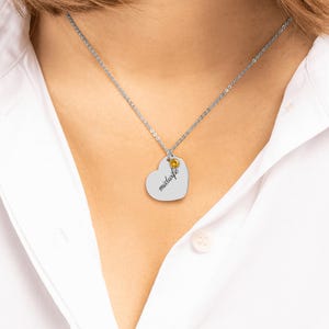 May include: A silver-toned necklace featuring a heart-shaped pendant engraved with the word midwife in a cursive script. The pendant is accented with a small, round, yellow-gold colored gemstone set near the top of the heart.