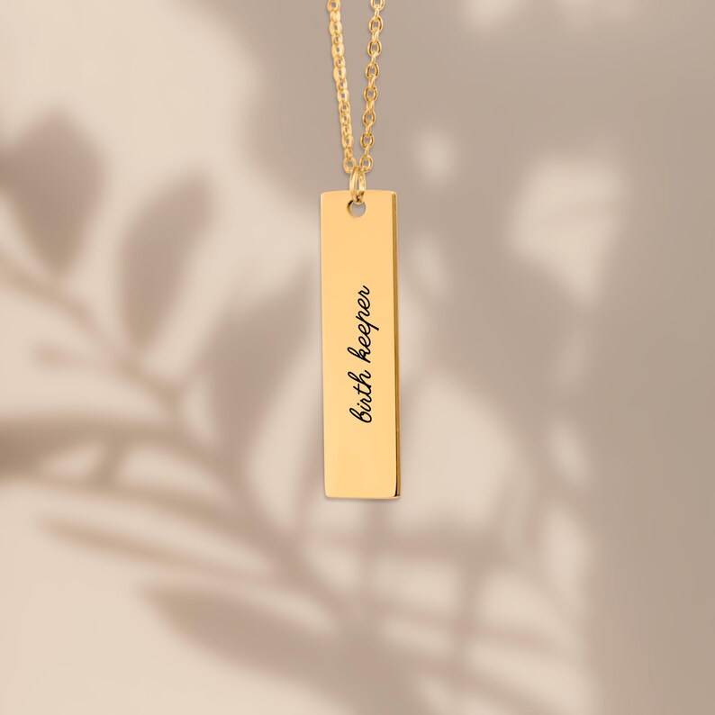 Op de afbeelding: Een goudkleurige ketting met een verticale staafhanger, een fijne ketting en een rechthoekig metalen plaatje. Het plaatje is gegraveerd met de zwarte cursieve tekst "birth keeper", verticaal gecentreerd op het gladde oppervlak.