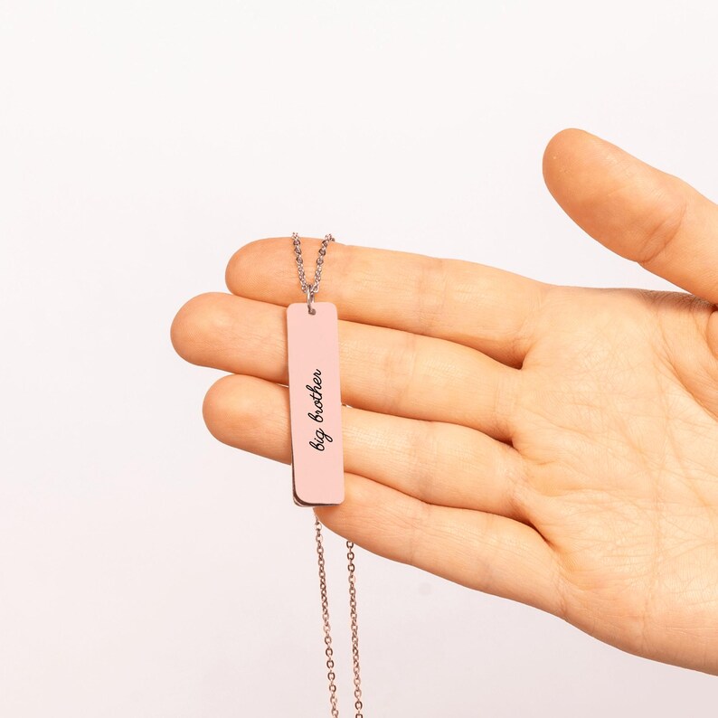 Puede incluir: Un collar con colgante de barra vertical en tono oro rosa que presenta el texto grabado big brother en una fuente cursiva. La barra met&aacute;lica rectangular cuelga de una cadena fina a juego con un acabado pulido y liso.