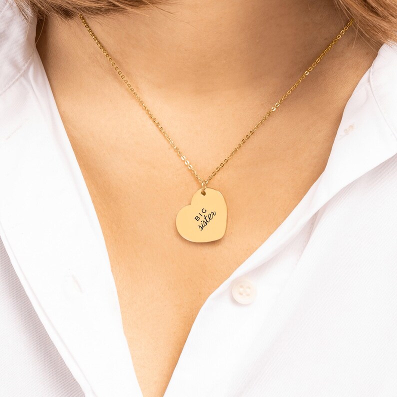 May include: A gold-toned necklace featuring a heart-shaped pendant with the text BIG sister engraved in black. The pendant hangs from a delicate gold-toned chain, resting against a white collared shirt.