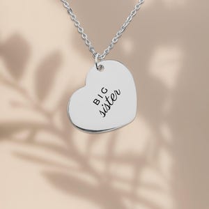 May include: A silver-toned heart-shaped pendant necklace featuring the engraved text "BIG sister" in a combination of block and script lettering. The pendant hangs from a delicate silver-toned chain, creating a simple and polished jewelry piece.
