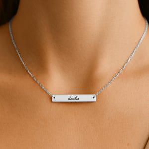 Pode incluir: Um colar de barra prateado com uma corrente fina e um pingente retangular horizontal. O pingente tem gravada a palavra doula em uma fonte cursiva preta, centralizada na superf&iacute;cie met&aacute;lica lisa e polida do objeto.